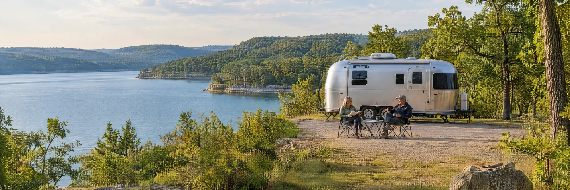 Illustration of campers enjoying relaxing at camp beside a rounded silver travel trailer in a fictional camping setting inspired by Mountain Home, AR.