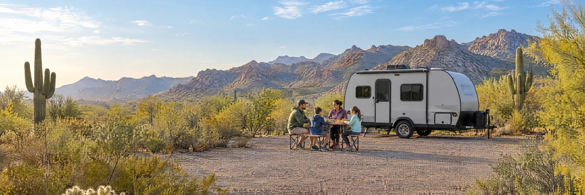 Illustration of campers enjoying a family picnic beside a compact travel trailer in a fictional camping setting inspired by Surprise, AZ.