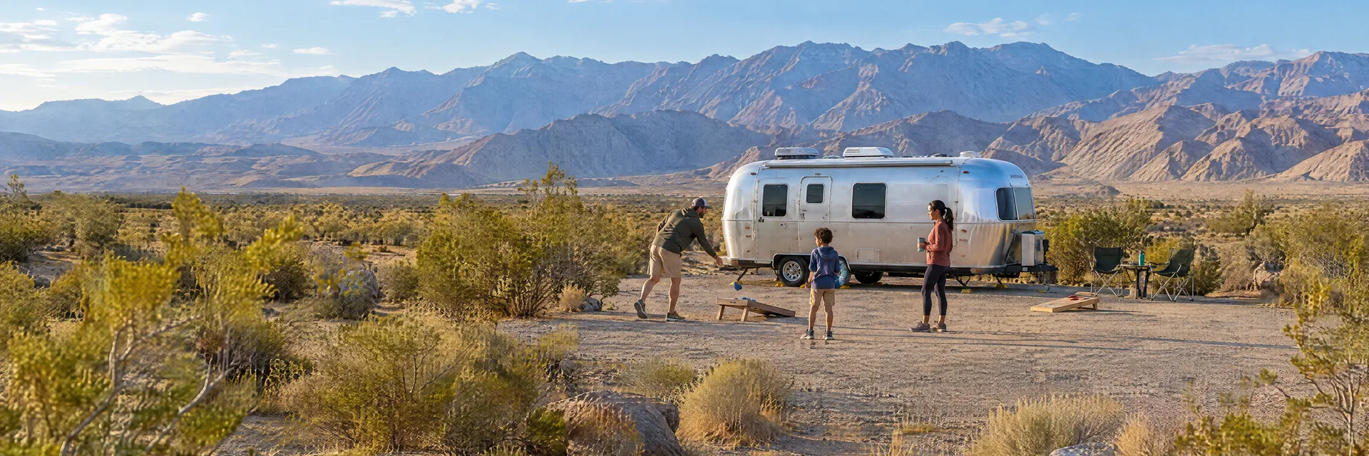 Illustration of campers enjoying family campsite activities beside a rounded silver travel trailer in a fictional camping setting inspired by Desert Hot Springs, CA.