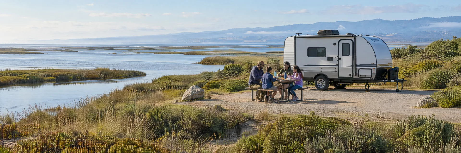 Illustration of campers enjoying a family picnic beside a compact travel trailer in a fictional camping setting inspired by Moss Landing, CA.