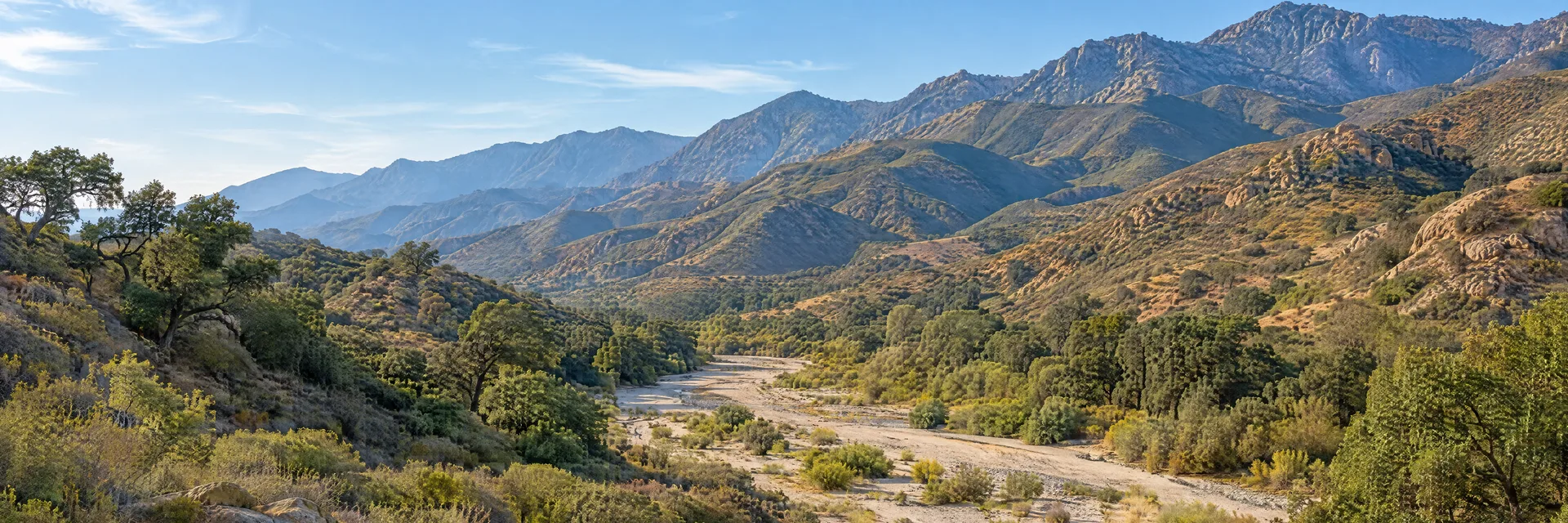 Illustration inspired by Sunland, CA: southern California foothill country, dry chaparral slopes, scattered live oaks and a broad sandy creek wash below rugged mountain ridges.