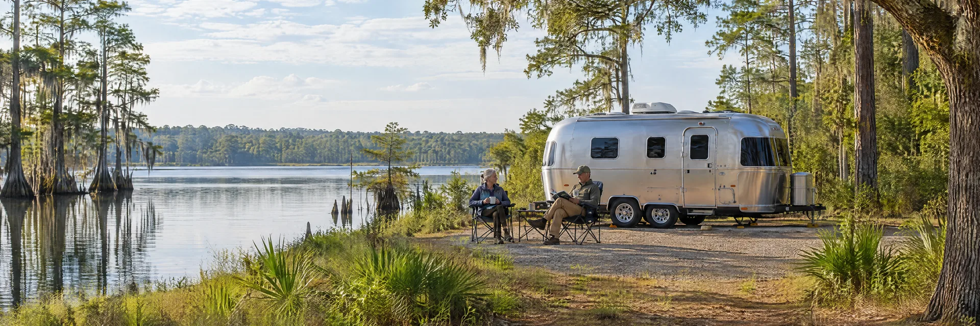 Illustration of campers enjoying relaxing at camp beside a rounded silver travel trailer in a fictional camping setting inspired by Ft. McCoy, FL.