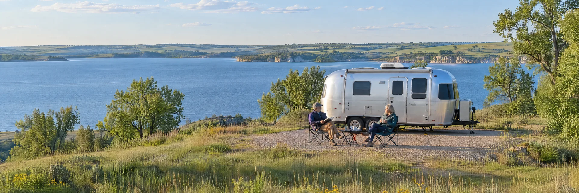 Illustration of campers enjoying relaxing at camp beside a rounded silver travel trailer in a fictional camping setting inspired by Junction City, KS.