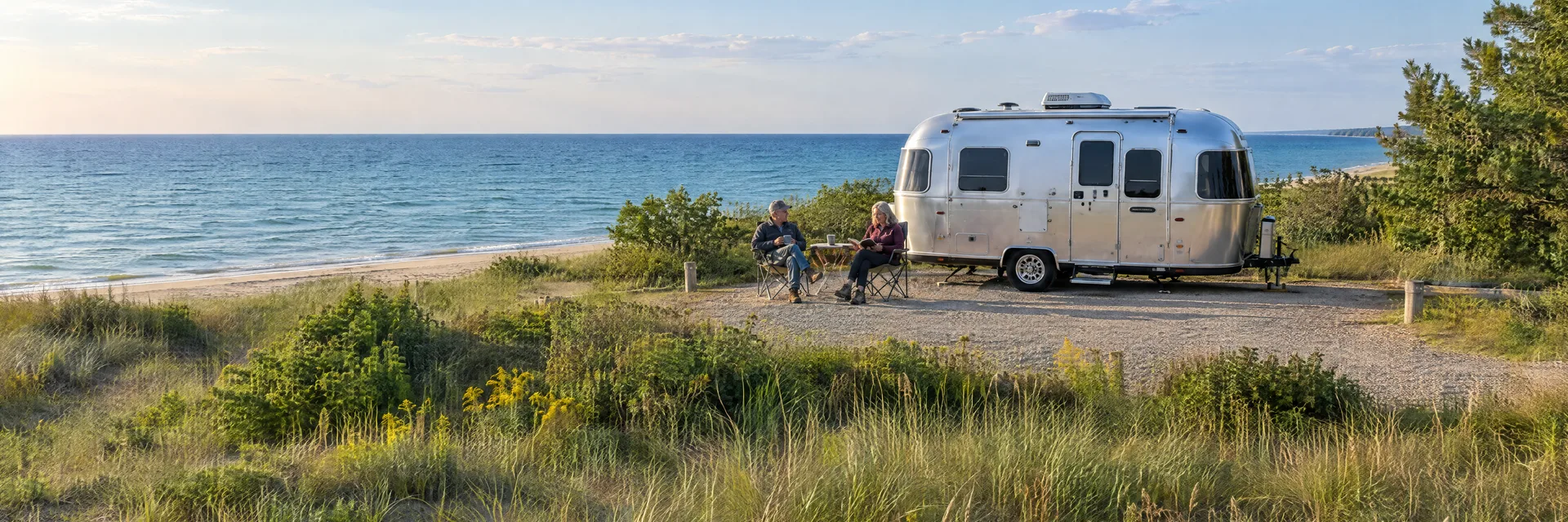 Illustration of campers enjoying relaxing at camp beside a rounded silver travel trailer in a fictional camping setting inspired by Benton Harbor, MI.