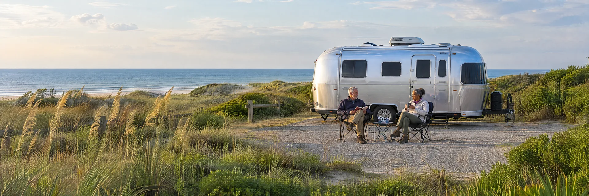 Illustration of campers enjoying relaxing at camp beside a rounded silver travel trailer in a fictional camping setting inspired by Buxton, NC.