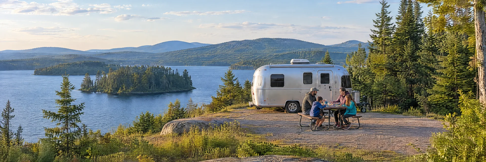 Illustration of campers enjoying a family picnic beside a rounded silver travel trailer in a fictional camping setting inspired by Cranberry Lake, NY.