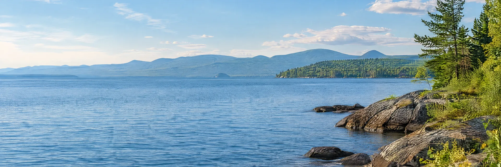 Illustration inspired by PLATTSBURGH, NY: lake Champlain-inspired open blue water, rocky shore and distant wooded mountains.