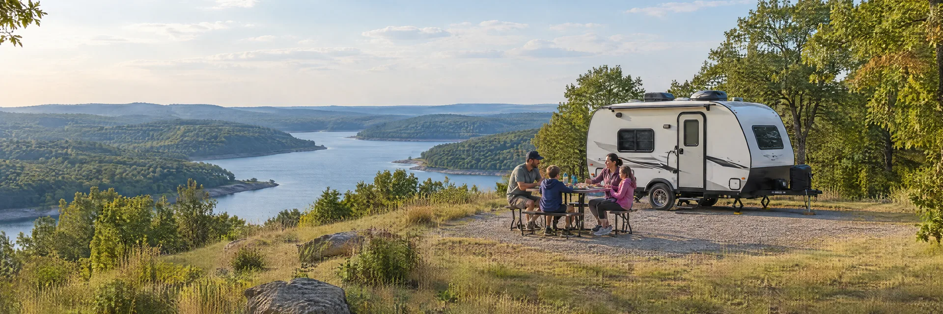 Illustration of campers enjoying a family picnic beside a compact travel trailer in a fictional camping setting inspired by Claremore, OK.