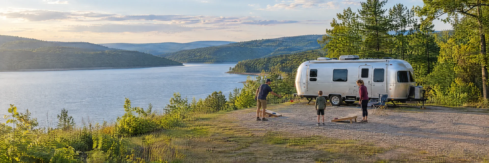 Illustration of campers enjoying family campsite activities beside a rounded silver travel trailer in a fictional camping setting inspired by Huntingdon, PA.