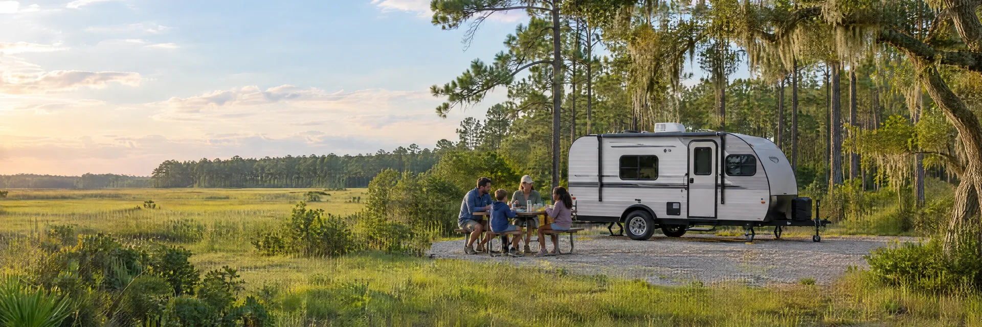 Illustration of campers enjoying a family picnic beside a compact travel trailer in a fictional camping setting inspired by Bowman, SC.