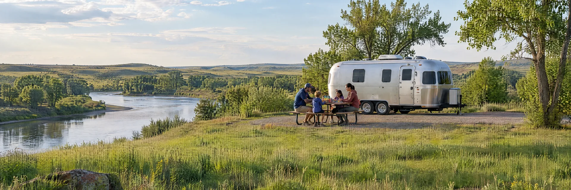 Illustration of campers enjoying a family picnic beside a rounded silver travel trailer in a fictional camping setting inspired by Sioux Falls, SD.