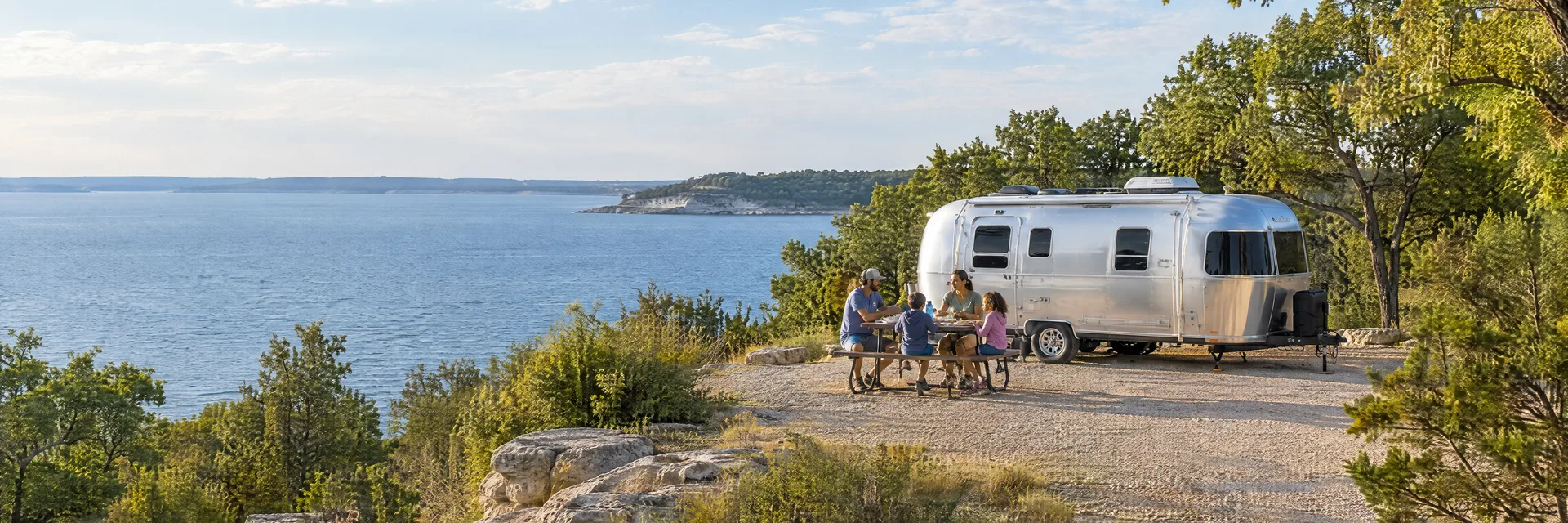 Illustration of campers enjoying a family picnic beside a rounded silver travel trailer in a fictional camping setting inspired by DENISON, TX.
