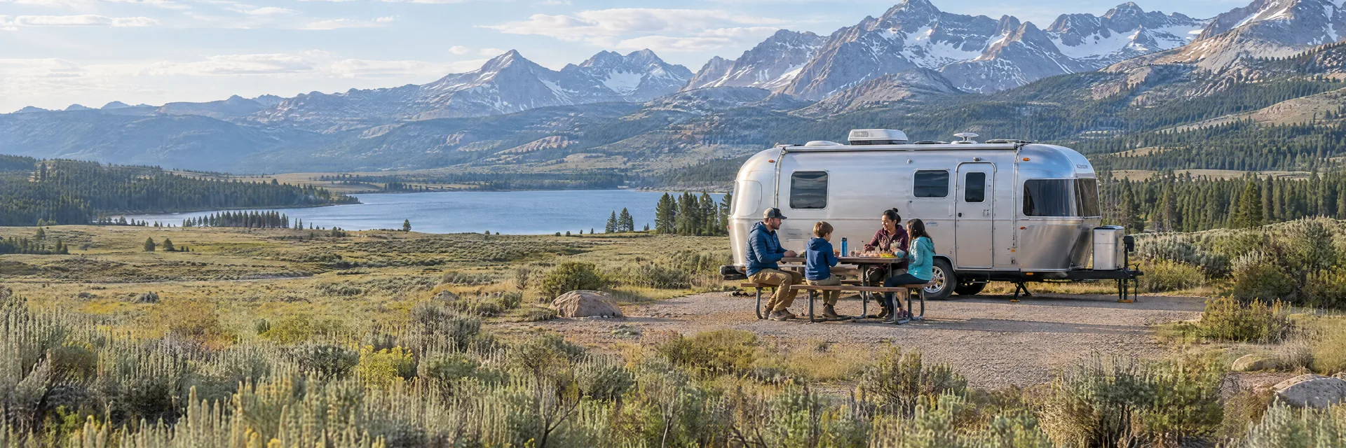 Illustration of campers enjoying a family picnic beside a rounded silver travel trailer in a fictional camping setting inspired by Pinedale, WY.