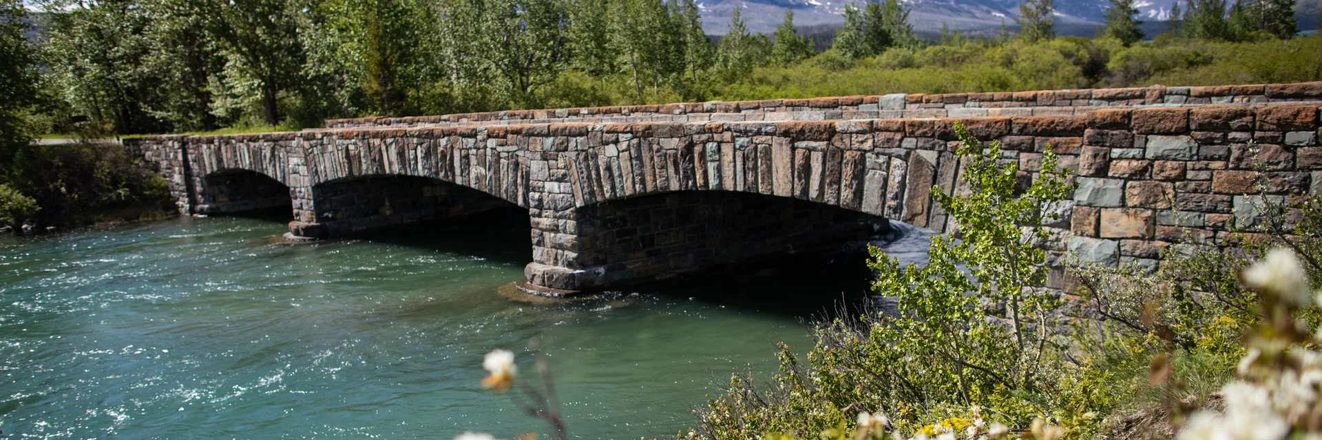 A historic stone bridge with forested slopes and mountains in Glacier National Park.