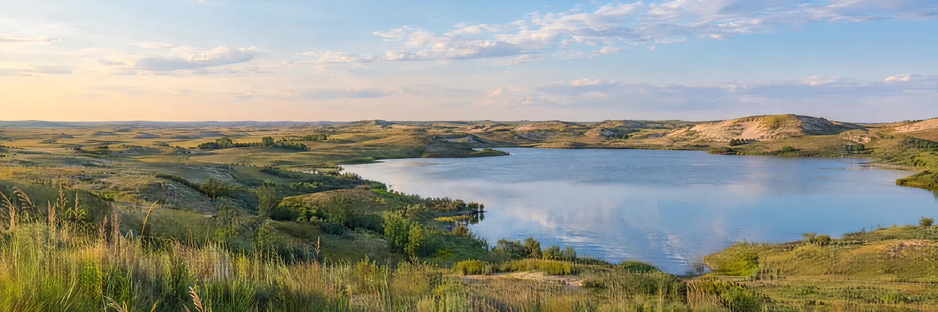 Illustration of representative natural scenery in Nebraska: sandhills grassland, gentle sandy ridges, small freshwater lake and wide sky.