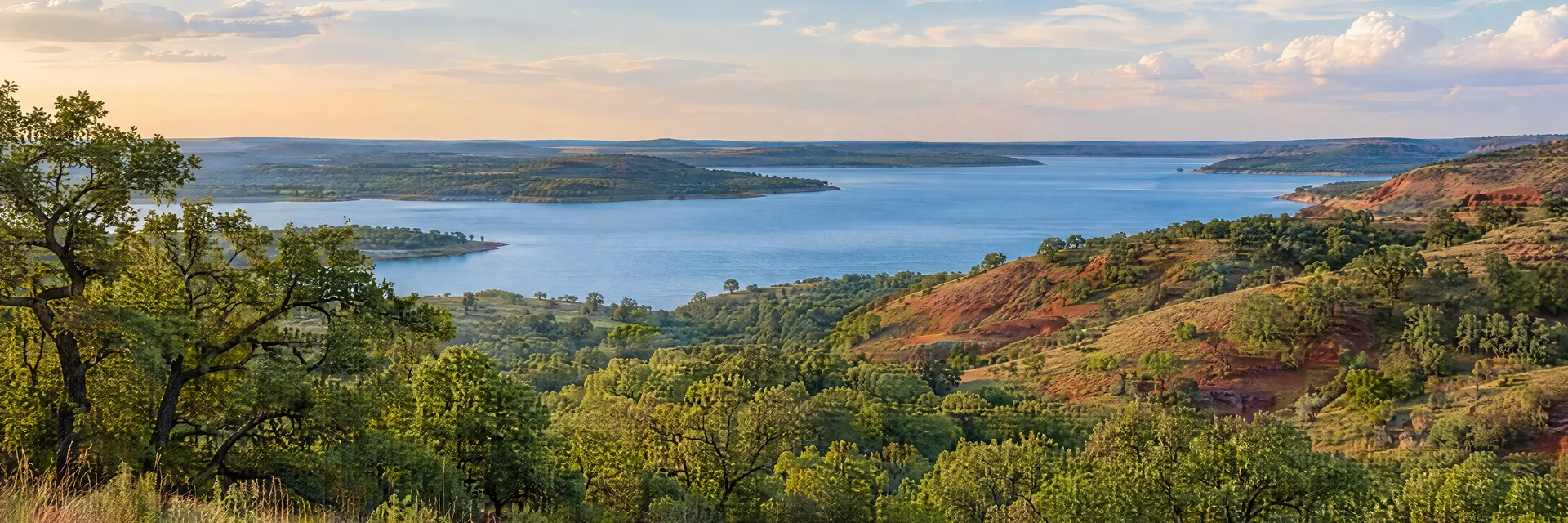 Illustration of representative natural scenery in Oklahoma: oak woodland, gently rolling red-earth hills and a broad freshwater reservoir.