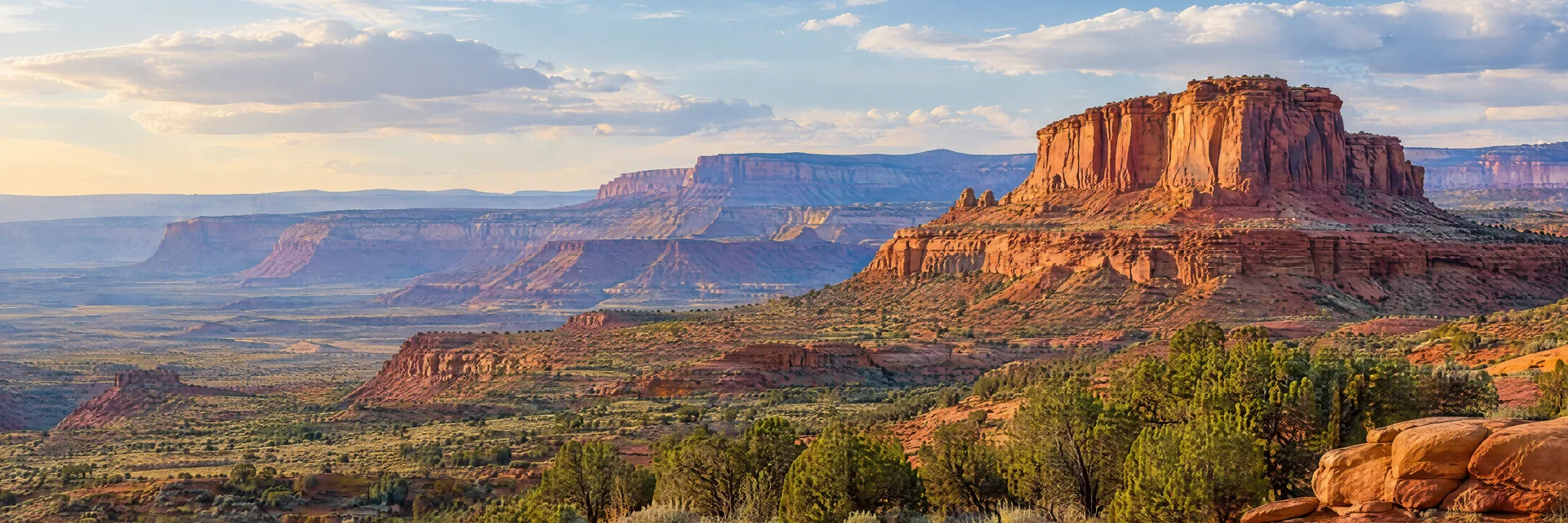 Illustration of representative natural scenery in Utah: red sandstone desert mesas, sparse native scrub and distant layered cliffs, no named arch.