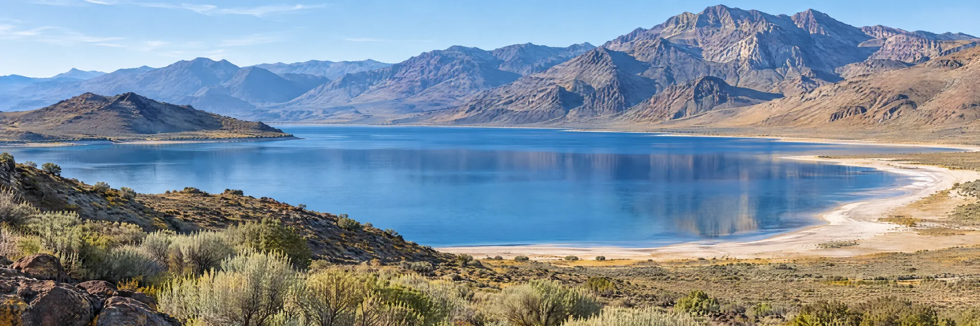 AI-generated illustration inspired by Nevada: Great Basin desert landscape, sagebrush and a broad blue desert lake edged by pale sandy shoreline, rugged barren mountains.
